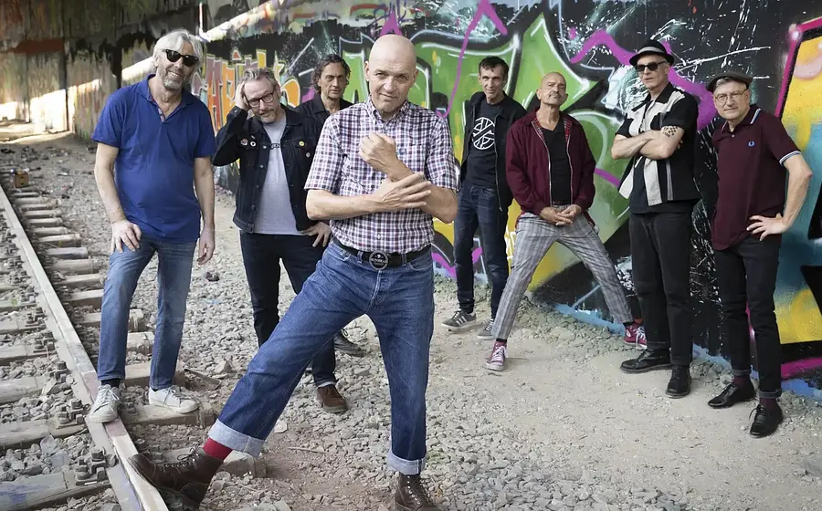 Foto zum 36 Jahre Backstage Jubiläum mit acht Personen in Freizeitkleidung vor einer bunt besprühten Graffitiwand entlang von Bahngleisen, die Gruppe steht verteilt zwischen Schotterbett und Wand, eine Person im karierten Kurzarmhemd posiert im Vordergrund.