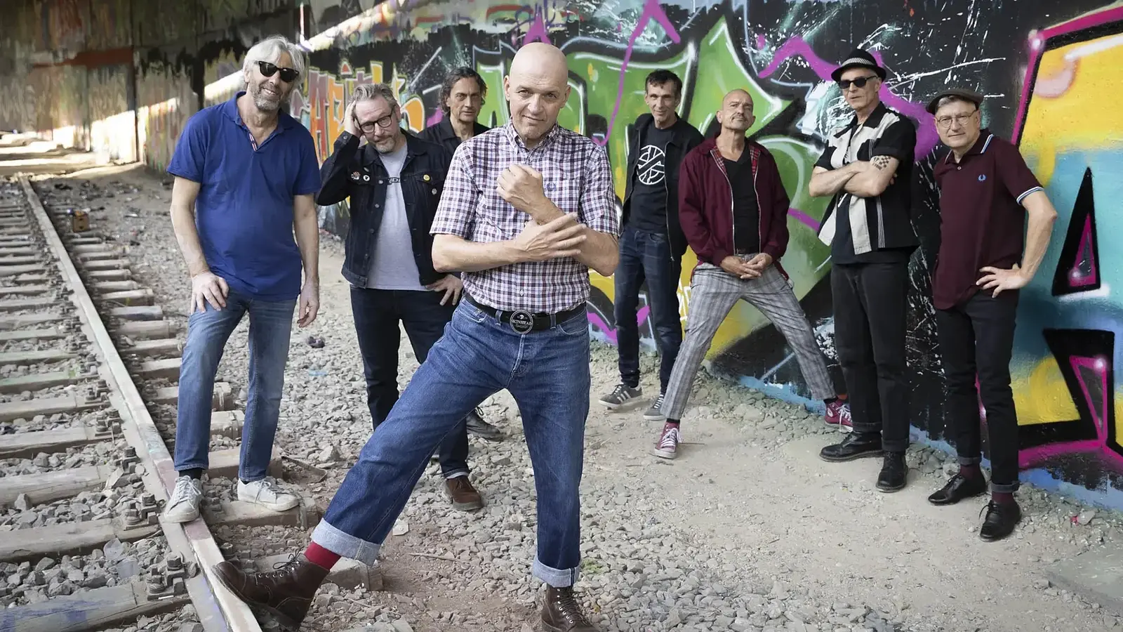 Foto zum 36 Jahre Backstage Jubiläum mit acht Personen in Freizeitkleidung vor einer bunt besprühten Graffitiwand entlang von Bahngleisen, die Gruppe steht verteilt zwischen Schotterbett und Wand, eine Person im karierten Kurzarmhemd posiert im Vordergrund.