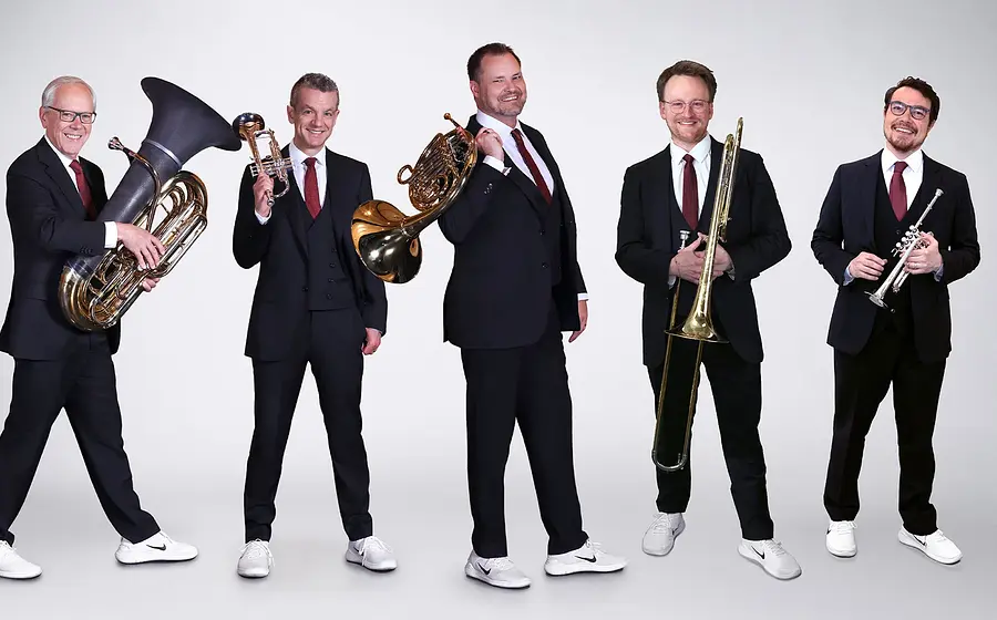 Canadian Brass mit fünf Musikern in dunklen Anzügen und weißen Turnschuhen vor hellem Hintergrund, jeweils mit Blechblasinstrumenten wie Tuba, Trompete, Horn und Posaune in stehender Studioaufnahme.