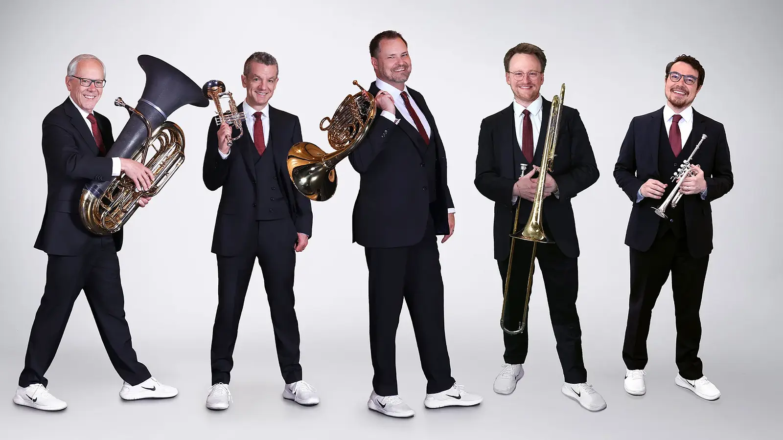 Canadian Brass mit fünf Musikern in dunklen Anzügen und weißen Turnschuhen vor hellem Hintergrund, jeweils mit Blechblasinstrumenten wie Tuba, Trompete, Horn und Posaune in stehender Studioaufnahme.