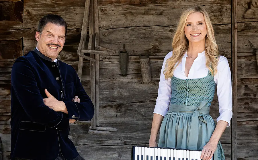 Eine Dame mit Instrument und ein Herr, beide in Tracht vor hölzernem Hintergrund