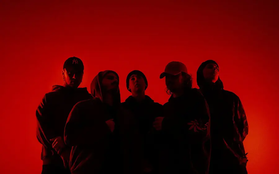 Bandfoto von Accvsed mit fünf Bandmitgliedern in dunkler Kleidung vor leuchtend rotem Hintergrund, darunter Personen mit Kapuzenpullovern, Mützen und Kappen in einer dicht stehenden Gruppenformation.