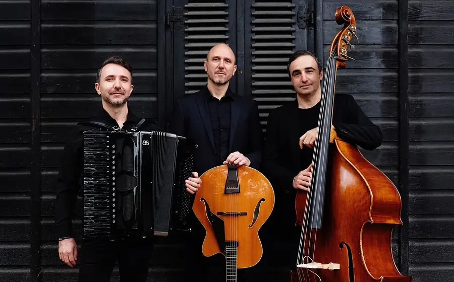 Die Band Acoustic Caravan vor einer schwarzen Holzwand. Drei Musiker in schwarzer Kleidung mit Akkordeon, Gitarre und Kontrabass. Die Instrumente setzen braune und schwarze Farbakzente.