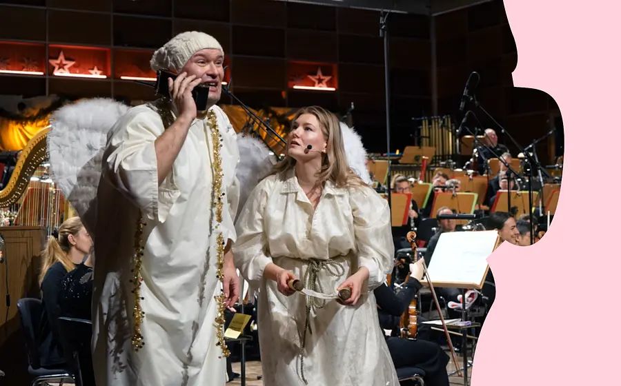 Zwei als Engel verkleidete Personen stehen vor einem Orchester auf der Bühne; eine Person hält ein Telefon. Im Hintergrund Musikerinnen und Musiker sowie eine Harfe.