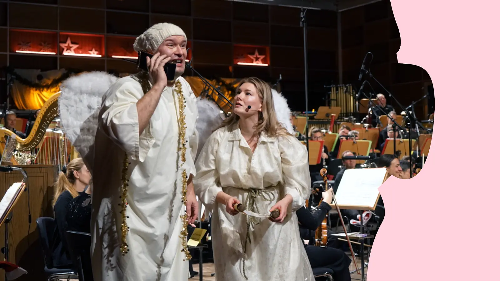 Zwei als Engel verkleidete Personen stehen vor einem Orchester auf der Bühne; eine Person hält ein Telefon. Im Hintergrund Musikerinnen und Musiker sowie eine Harfe.
