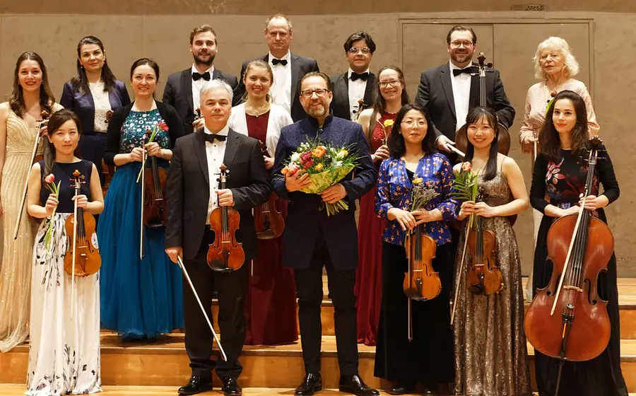 Ein Gruppenfoto des festlich gekleidetetn orchesters mit Instrumenten sowie einer Person ind er Mitte mit Blumenstrauß