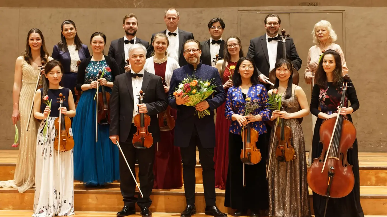 Ein Gruppenfoto des festlich gekleidetetn orchesters mit Instrumenten sowie einer Person ind er Mitte mit Blumenstrauß