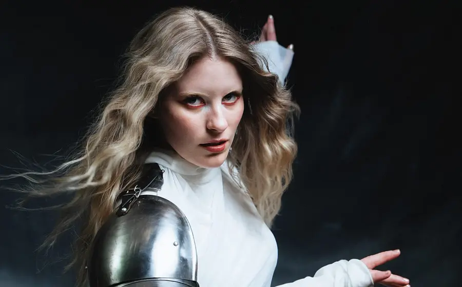 Person mit blondem, offenem Haar trägt ein weißes Oberteil und eine metallene Schulterrüstung und posiert vor dunklem Hintergrund.