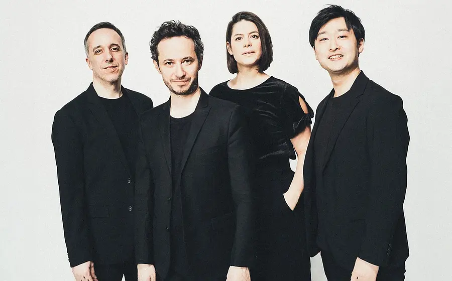 Quatuor Ébène mit vier Musiker:innen in schwarzer Kleidung vor hellem Hintergrund, nebeneinander stehend in Studioaufnahme ohne sichtbare Instrumente.