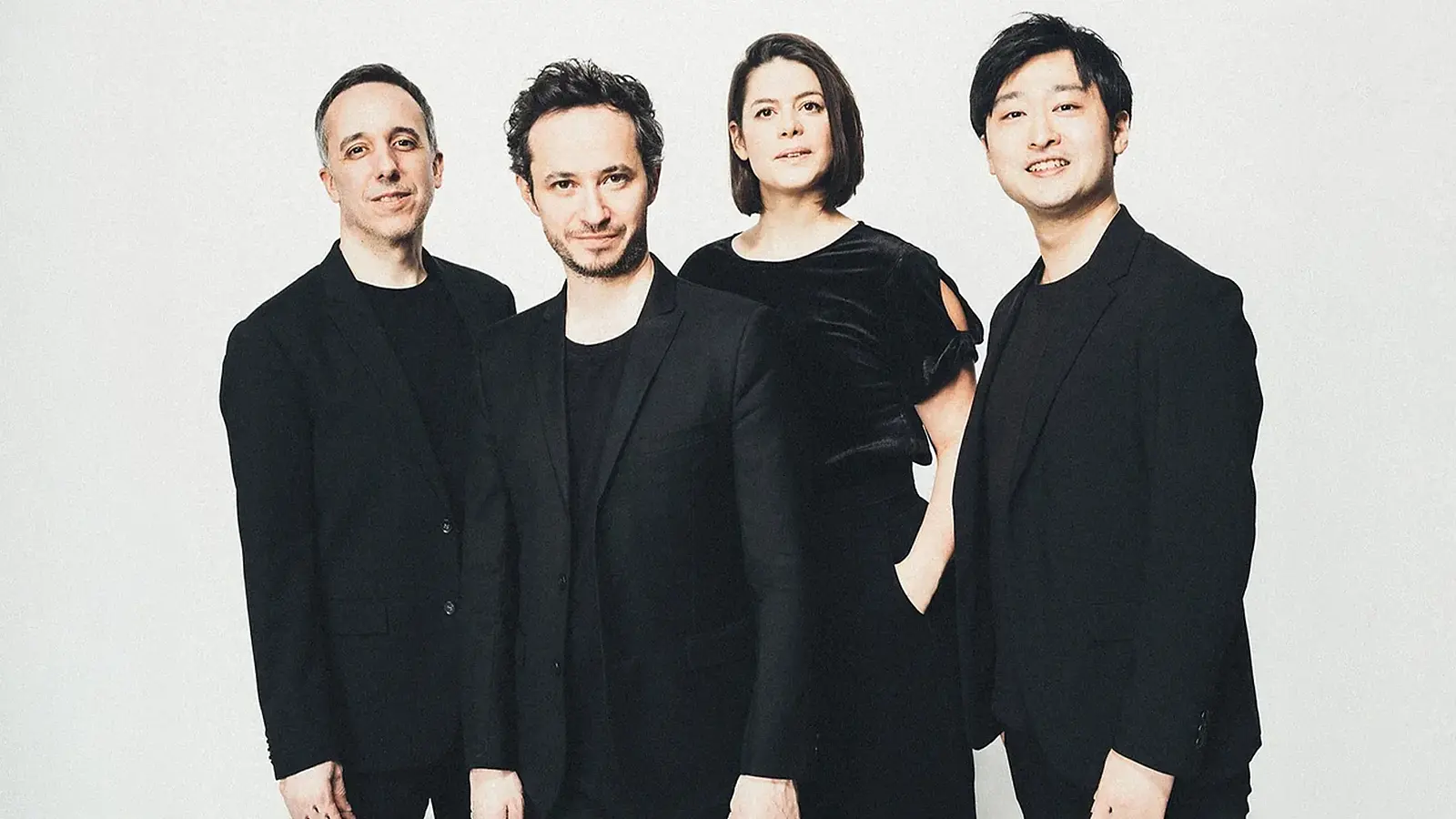 Quatuor Ébène mit vier Musiker:innen in schwarzer Kleidung vor hellem Hintergrund, nebeneinander stehend in Studioaufnahme ohne sichtbare Instrumente.