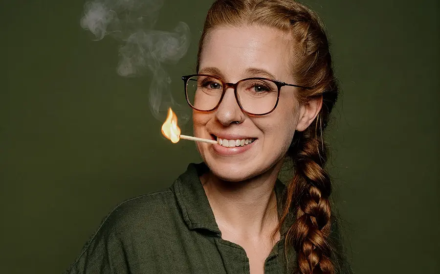 Sara Brandhuber in grüner Bluse vor grünem Hintergrund lächelnd mit einem brennenden Streichholz zwischen den Zähnen