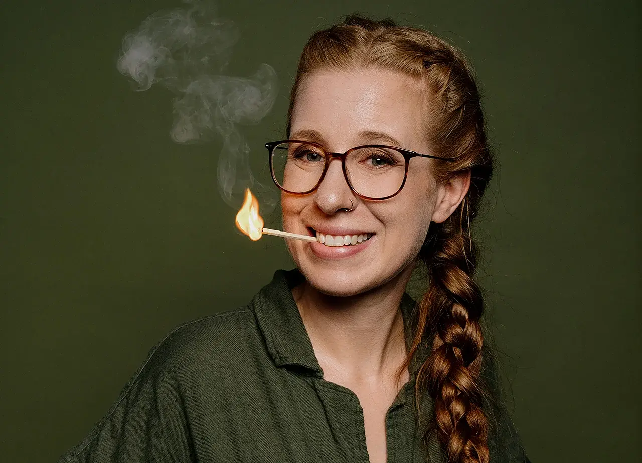 Sara Brandhuber in grüner Bluse vor grünem Hintergrund lächelnd mit einem brennenden Streichholz zwischen den Zähnen