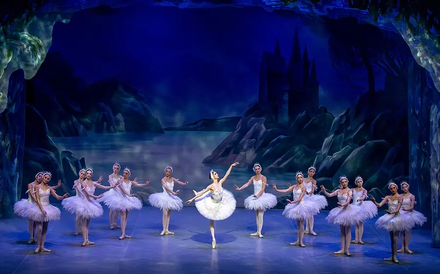 Bühnenfoto aus Schwanensee mit einer Solotänzerin in weißem Tutu im Zentrum, umgeben von weiteren Balletttänzerinnen in weißen Kostümen auf einer blau beleuchteten Bühne vor einer nächtlichen Seen- und Schlosskulisse