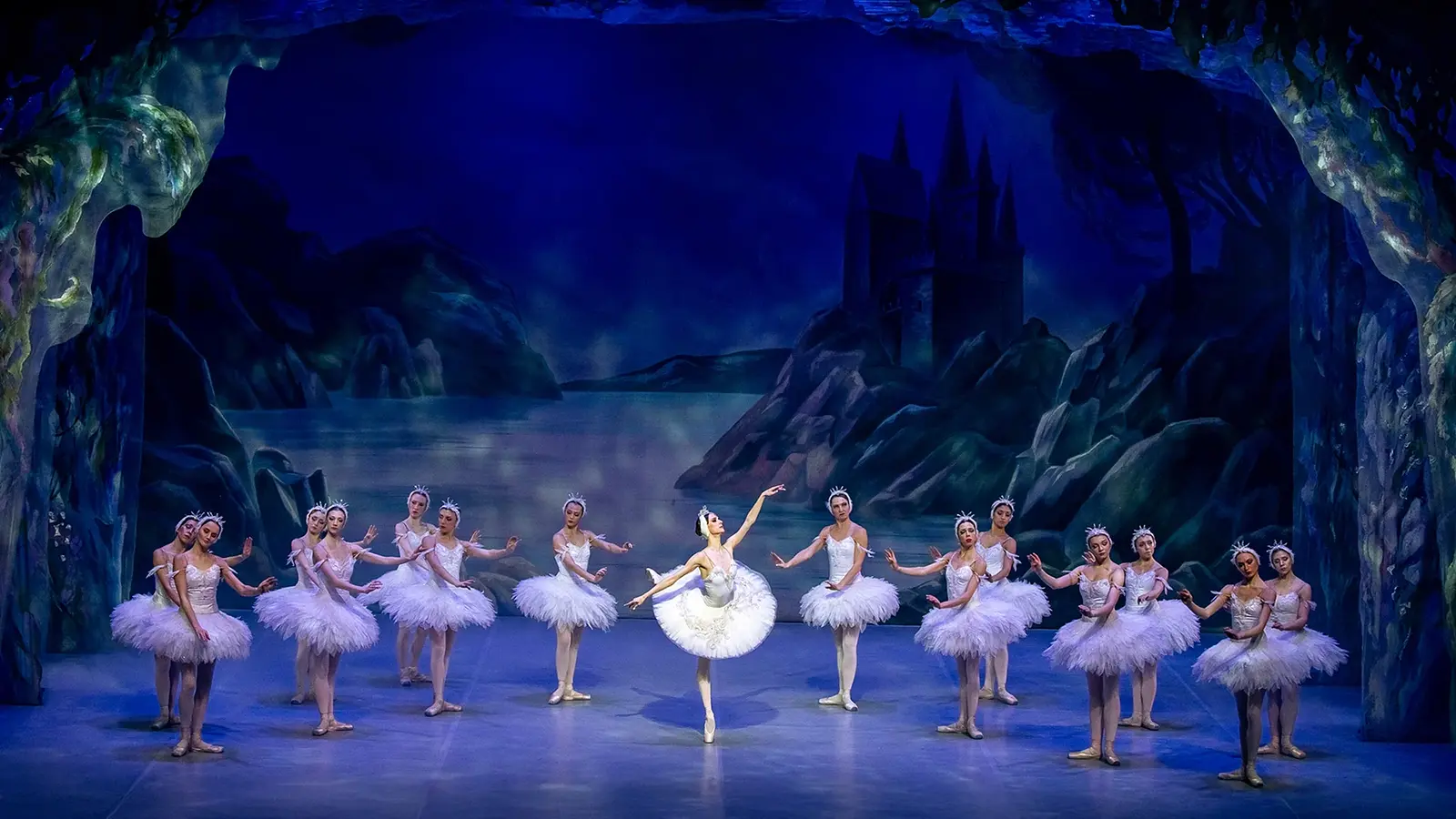 Bühnenfoto aus Schwanensee mit einer Solotänzerin in weißem Tutu im Zentrum, umgeben von weiteren Balletttänzerinnen in weißen Kostümen auf einer blau beleuchteten Bühne vor einer nächtlichen Seen- und Schlosskulisse