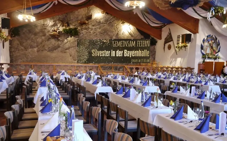 Ein traditionell bayerisch geschmückter Festsaal mit festlich gedeckten Tischen in Reihen und Veranstaltungstitel im Hintergrund