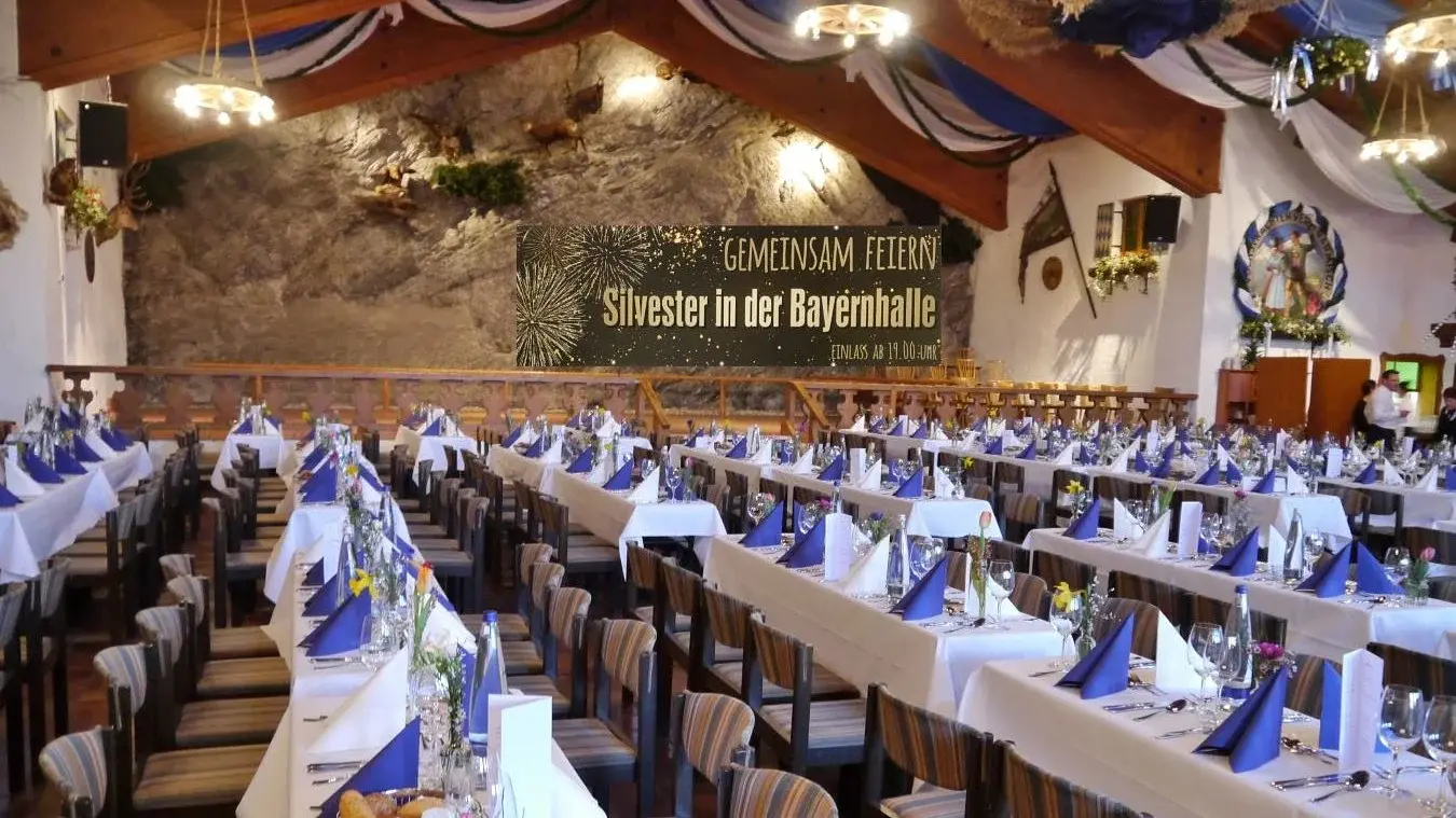 Ein traditionell bayerisch geschmückter Festsaal mit festlich gedeckten Tischen in Reihen und Veranstaltungstitel im Hintergrund