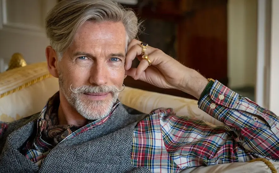 Porträtfoto einer Person mit grauem Haar und gezwirbeltem Schnurrbart, in kariertem Hemd, grauer Weste und Halstuch, auf einem hellen Sofa in einem eleganten Innenraum sitzend
