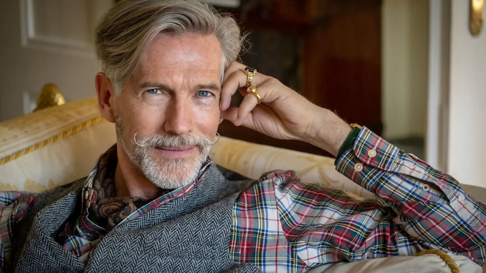 Porträtfoto einer Person mit grauem Haar und gezwirbeltem Schnurrbart, in kariertem Hemd, grauer Weste und Halstuch, auf einem hellen Sofa in einem eleganten Innenraum sitzend