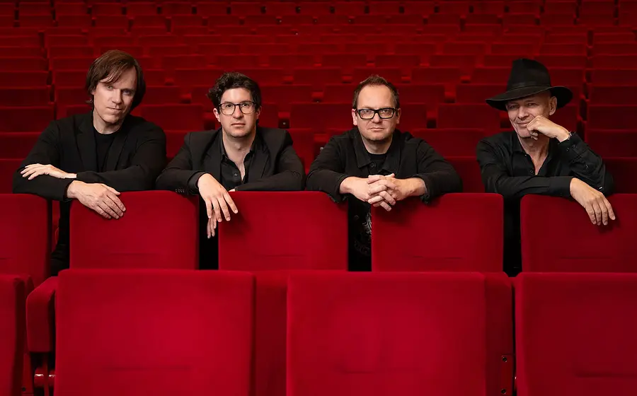 Pressefoto von Vokal Total Rock 4 mit vier Musikern in schwarzer Kleidung, die sich über die Rückenlehnen roter Kinosessel lehnen, vor einem leeren Zuschauerraum mit Reihen roter Sitze.