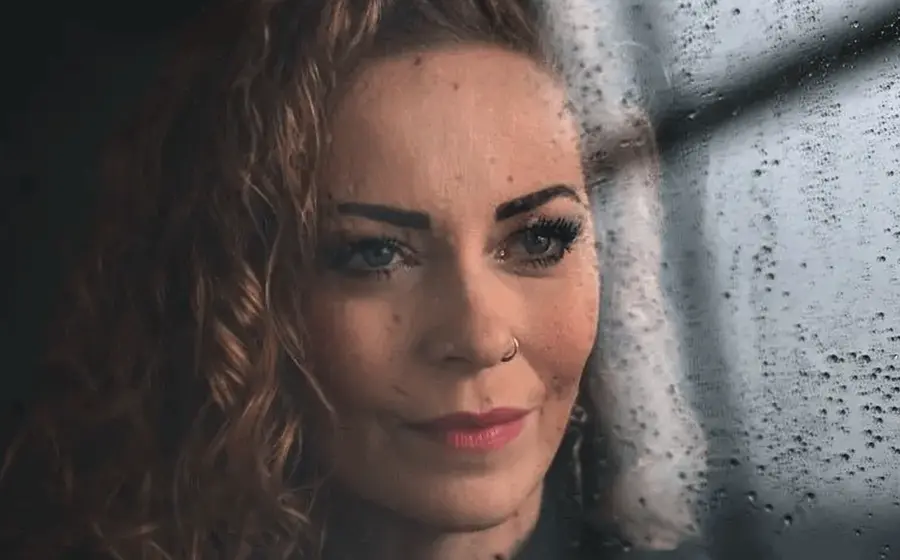 Porträt einer Person mit lockigem Haar und Nasenring hinter einer regennassen Fensterscheibe. Die Nahaufnahme zeigt den Blick durch die mit Wassertropfen bedeckte Scheibe vor unscharfem Hintergrund.