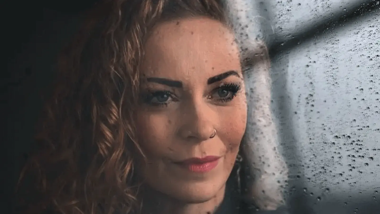 Porträt einer Person mit lockigem Haar und Nasenring hinter einer regennassen Fensterscheibe. Die Nahaufnahme zeigt den Blick durch die mit Wassertropfen bedeckte Scheibe vor unscharfem Hintergrund.