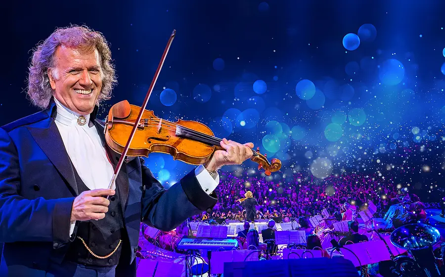 André Rieu steht im Vordergrund einer groß beleuchteten Konzertbühne und spielt eine Violine; im Hintergrund ist ein großes Orchester vor einem Publikum zu sehen, umgeben von blau‑violettem Licht und vielen schwebenden Lichtpunkten.
