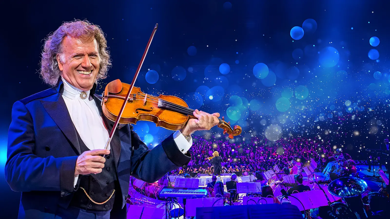 André Rieu steht im Vordergrund einer groß beleuchteten Konzertbühne und spielt eine Violine; im Hintergrund ist ein großes Orchester vor einem Publikum zu sehen, umgeben von blau‑violettem Licht und vielen schwebenden Lichtpunkten.