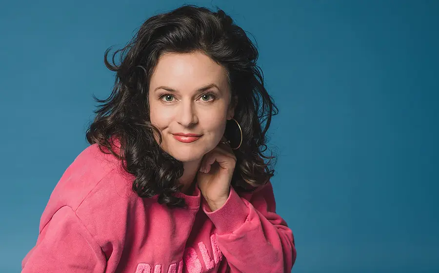 Angela Ascher in pinkem Sweatshirt lächelt in die Kamera, vor blauem Hintergrund
