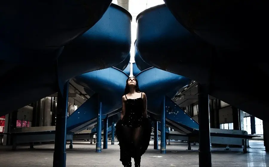 Foto einer Dame in dunkler Kleidung, stehend in einer industriellen Halle zwischen großen, blauen, trichterförmigen Metallstrukturen; symmetrische Perspektive mit Licht von oben.