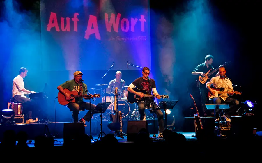 Liveauftritt der Band Auf A Wort auf einer Bühne mit farbiger Beleuchtung. Sechs Musiker spielen Gitarre, Bass, Schlagzeug und Keyboard, während im Hintergrund die Projektion Auf A Wort Die Songs von STS zu sehen ist.