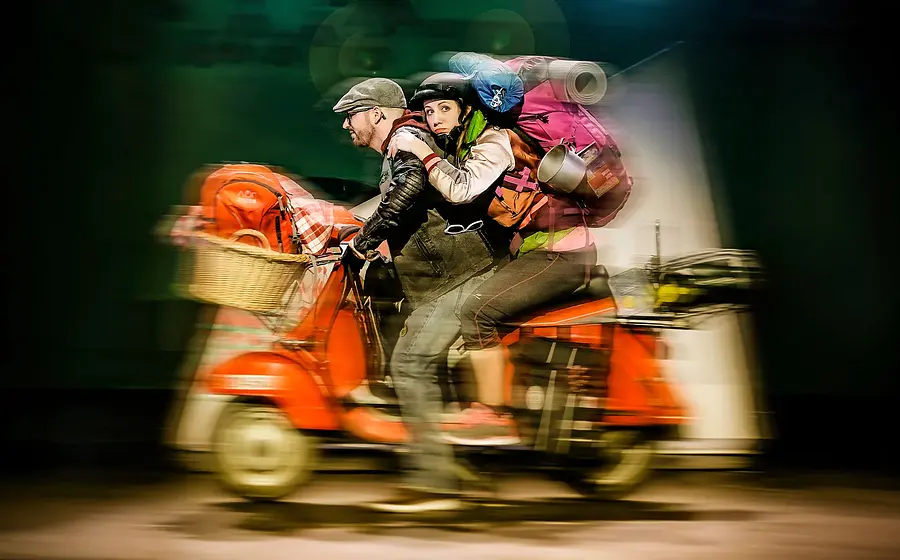 Zwei Personen fahren auf einem roten Roller mit Gepäck durch eine Szene, mit Bewegungsunschärfe und Bühnenlicht im Hintergrund.