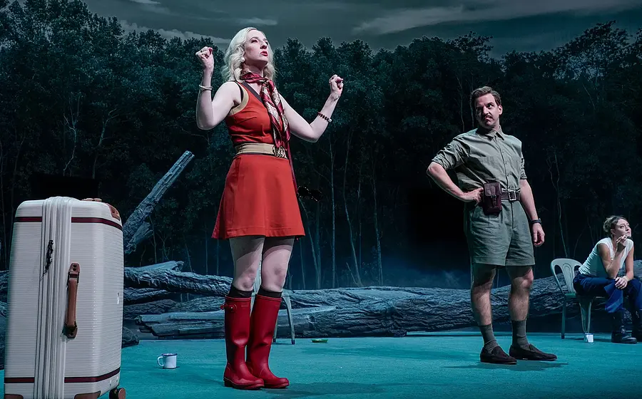Eine rot gekleidete Dame und ein Herr Stehen auf einer Bühne mit einem Wald im Hintergrund, ein Koffer vorne