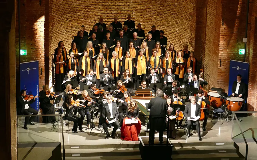 Bühnenfoto von Chor und Orchester bei einer Aufführung des Weihnachtsoratoriums in einem hohen Backsteinbau, mit Dirigent vor dem Ensemble, Solistinnen und Solisten im Vordergrund sowie Sängerinnen und Sängern mit goldfarbenen Schals auf gestaffelten Podesten im Hintergrund.