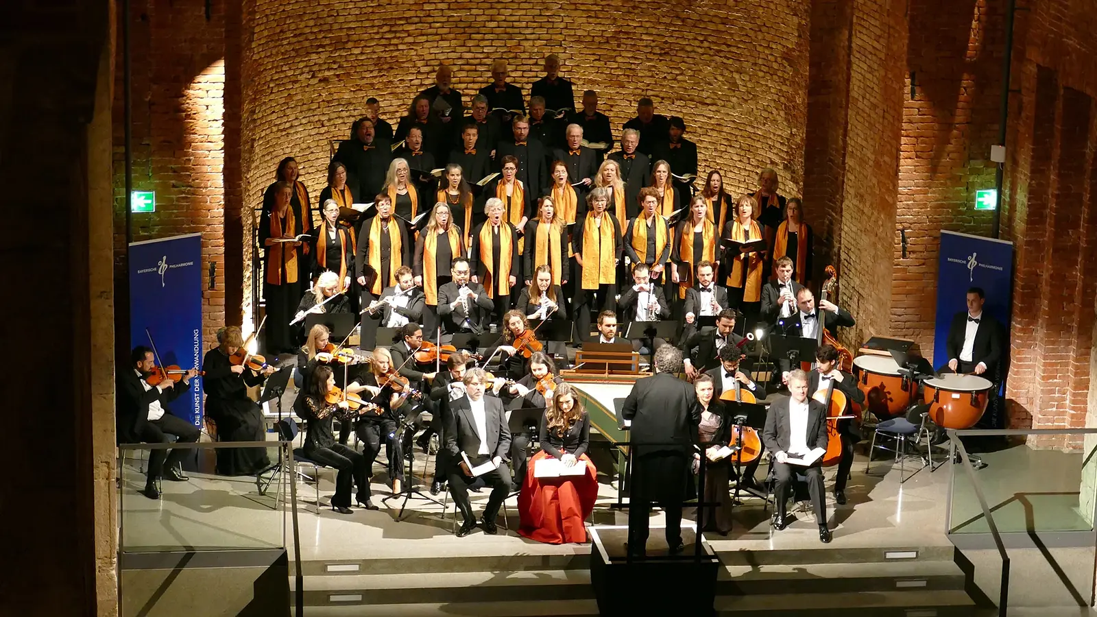 Bühnenfoto von Chor und Orchester bei einer Aufführung des Weihnachtsoratoriums in einem hohen Backsteinbau, mit Dirigent vor dem Ensemble, Solistinnen und Solisten im Vordergrund sowie Sängerinnen und Sängern mit goldfarbenen Schals auf gestaffelten Podesten im Hintergrund.