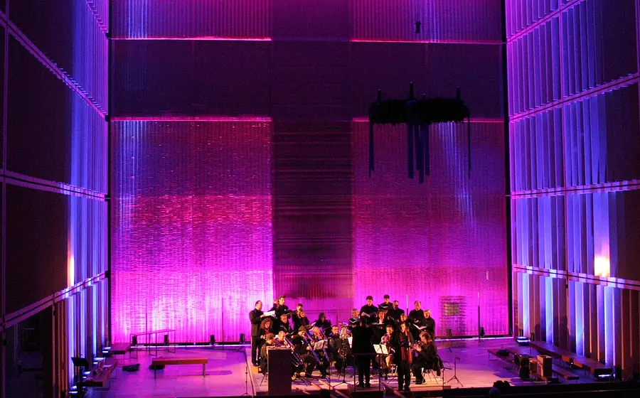 Bühnenfoto von Ascending Voices mit Chor und Kammerensemble auf einer Bühne der Herz-Jesu-Kirche, vor einer großflächig in Pink und Violett beleuchteten Architekturkulisse mit hohen Wandlamellen und transparentem Vorhang im Hintergrund