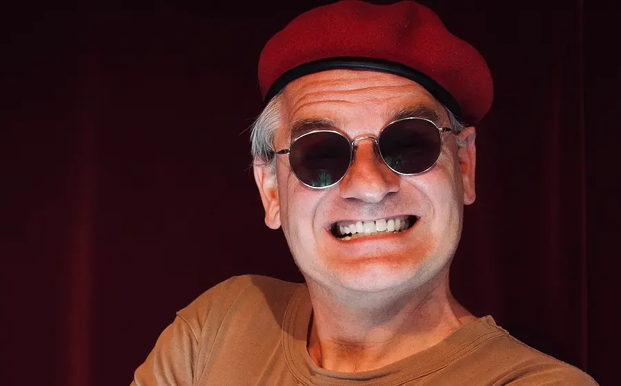 Person mit roter Baskenmütze und runder Sonnenbrille vor dunkelrotem Hintergrund. Die Person grinst weit, zeigt dabei ihre Zähne und trägt ein hellbraunes T Shirt.