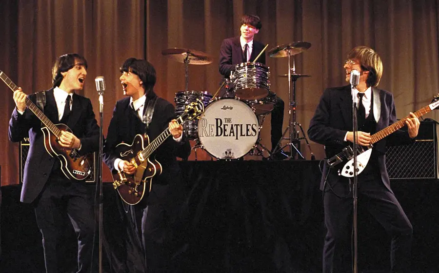 Bühnenfoto einer Beatles Tribute Band mit vier Musikern in schwarzen Anzügen und Pilzkopfperücken, drei Personen mit E Gitarren und eine am Schlagzeug, vor Mikrofonen und Verstärkern auf einer Bühne mit beigefarbenem Vorhang im Hintergrund.