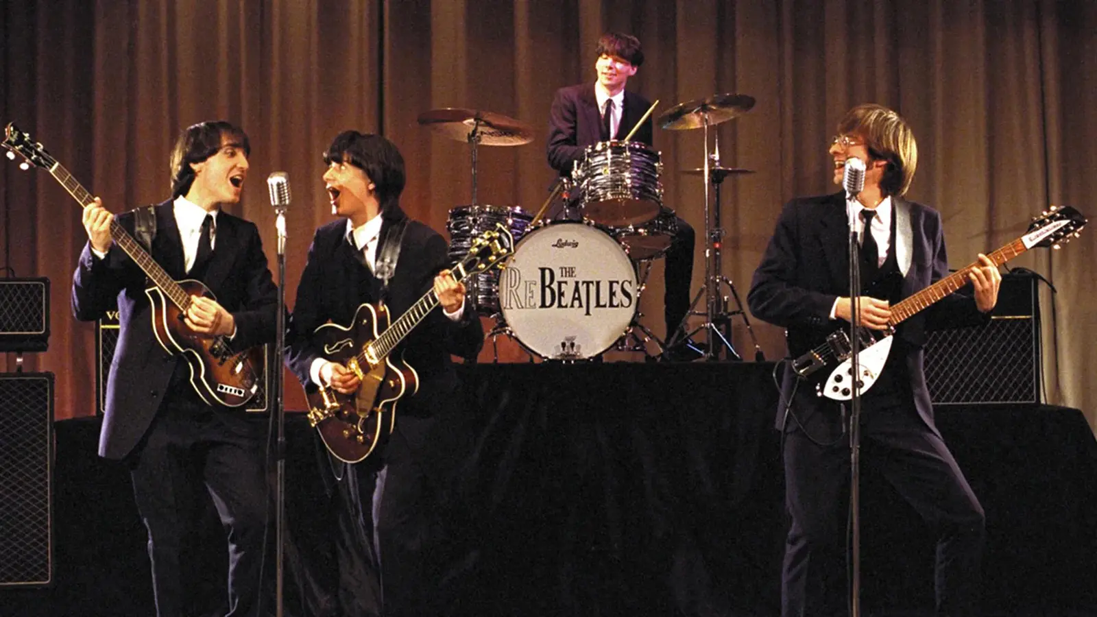 Bühnenfoto einer Beatles Tribute Band mit vier Musikern in schwarzen Anzügen und Pilzkopfperücken, drei Personen mit E Gitarren und eine am Schlagzeug, vor Mikrofonen und Verstärkern auf einer Bühne mit beigefarbenem Vorhang im Hintergrund.