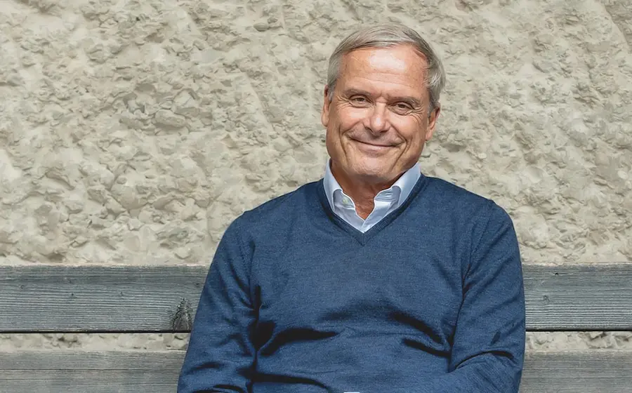 Axel Hacke lächelnd in blauem Pullover sitzt auf einer Holzbank vor einer groben Steinwand