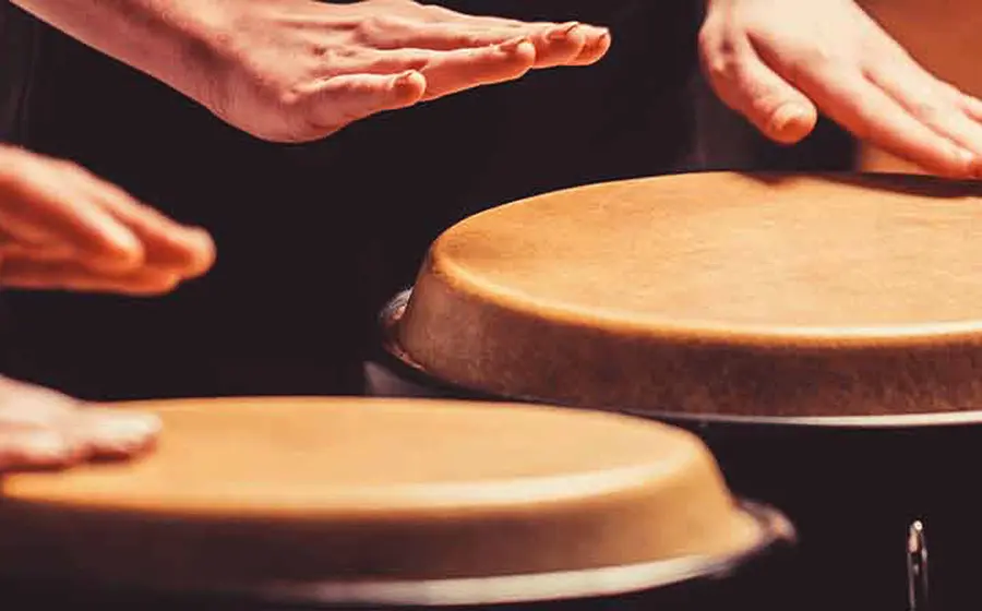 Nahaufnahme mehrerer Hände, die auf Conga Trommeln spielen. Die Aufnahme zeigt die Schlagbewegungen auf den naturfarbenen Trommelfellen vor dunklem Hintergrund.