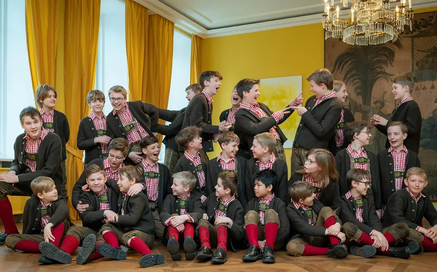 Kindergruppe in Tracht sitzt und spielt zusammen in einem hellen Raum mit gelben Vorhängen und Kronleuchter
