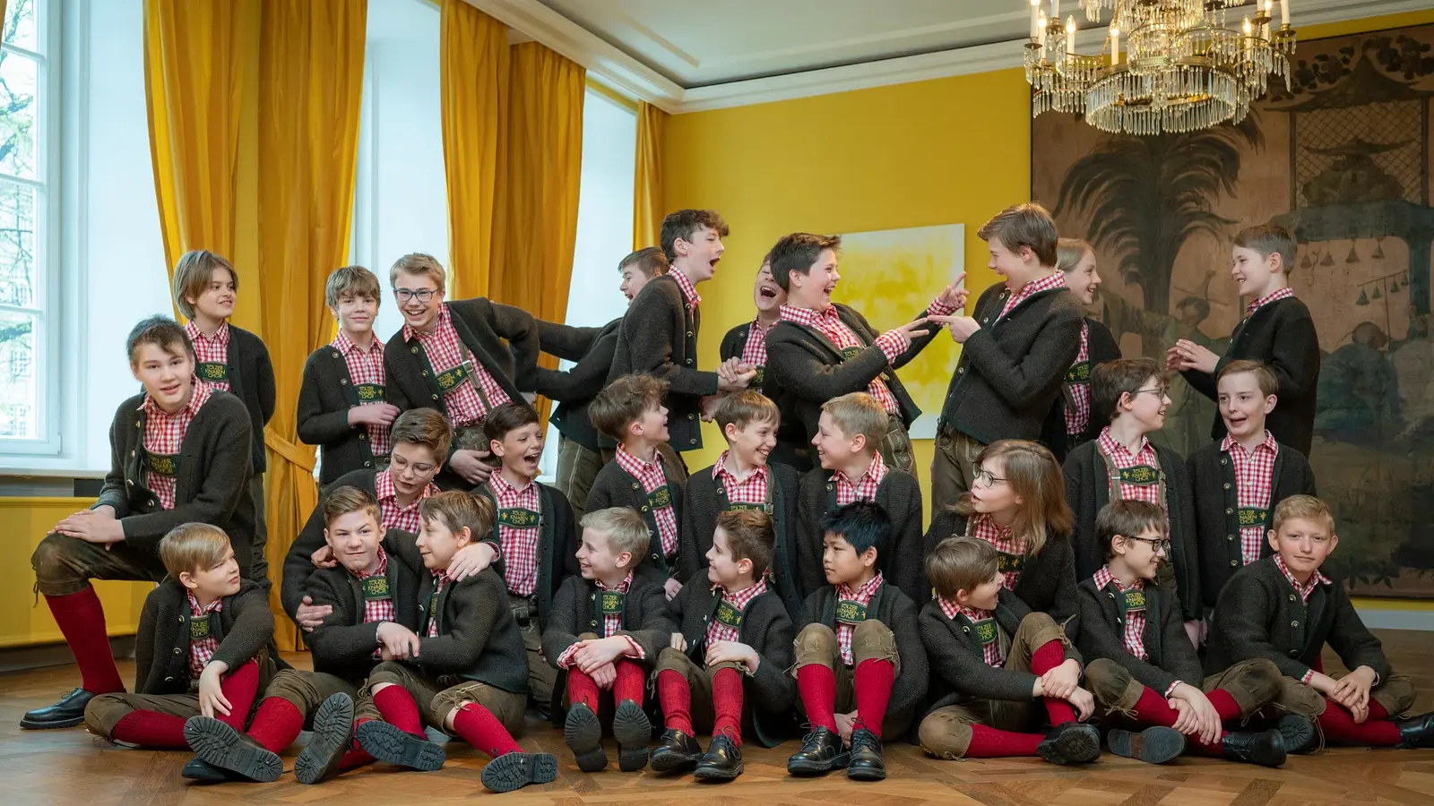 Kindergruppe in Tracht sitzt und spielt zusammen in einem hellen Raum mit gelben Vorhängen und Kronleuchter