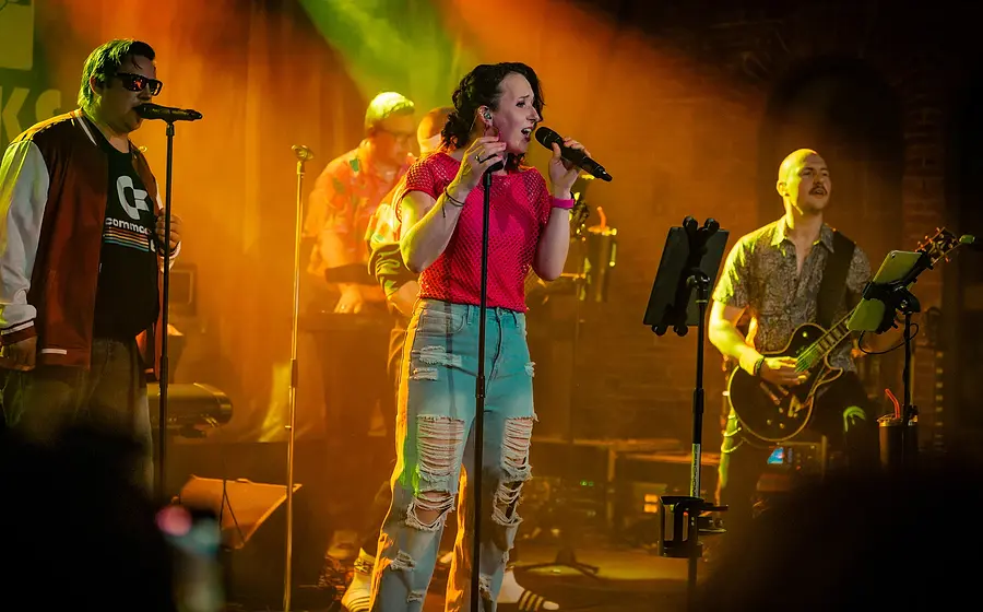 Band mit Sängerin, Sänger, Gitarrist und Keyboarder auf einer Bühne. Die Sängerin trägt ein pinkes Oberteil und hellblaue Jeans. Die Bühne ist in warmes orangefarbenes sowie grünes Licht getaucht, der Hintergrund bleibt dunkel.