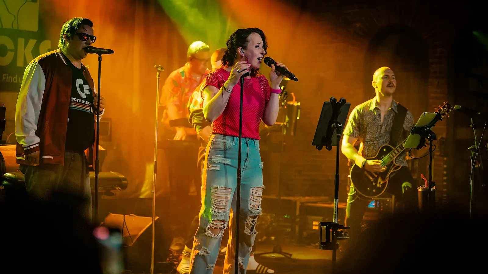 Band mit Sängerin, Sänger, Gitarrist und Keyboarder auf einer Bühne. Die Sängerin trägt ein pinkes Oberteil und hellblaue Jeans. Die Bühne ist in warmes orangefarbenes sowie grünes Licht getaucht, der Hintergrund bleibt dunkel.