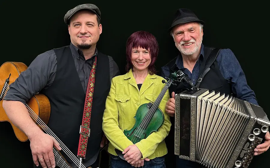 Bandfoto von Jodelwahnsinn mit drei Musikerinnen und Musikern vor schwarzem Hintergrund, mit Gitarre, Violine und Steirischer Harmonika, die mittlere Person in hellgrüner Jacke, die beiden anderen mit Hut und dunklen Westen.