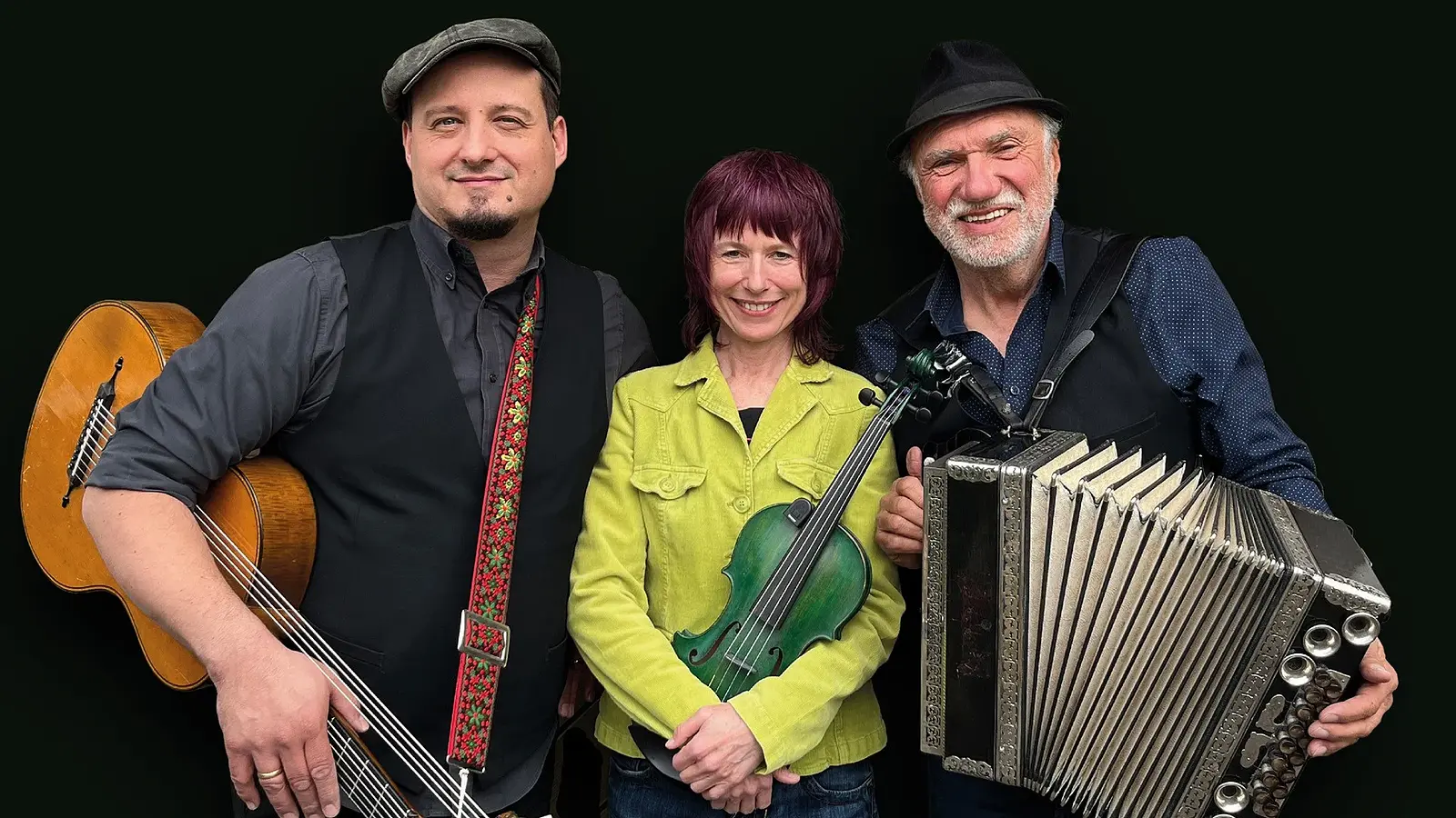 Bandfoto von Jodelwahnsinn mit drei Musikerinnen und Musikern vor schwarzem Hintergrund, mit Gitarre, Violine und Steirischer Harmonika, die mittlere Person in hellgrüner Jacke, die beiden anderen mit Hut und dunklen Westen.