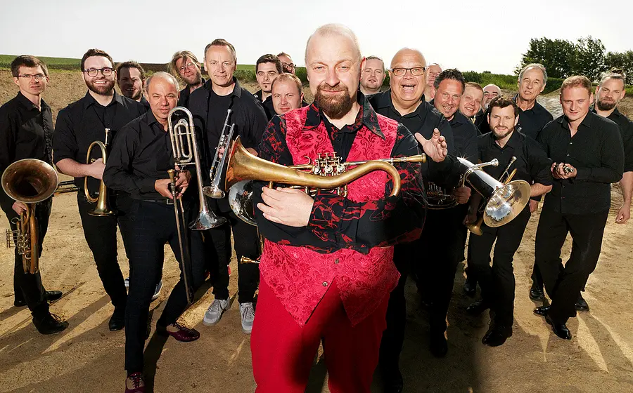 Gruppenfoto der Blasmusik Supergroup im Freien. Im Vordergrund hält eine Person in roter Weste ein Blasinstrument, während zahlreiche Musikerinnen und Musiker in schwarzer Kleidung mit Trompeten, Posaunen, Tuben und weiteren Blechblasinstrumenten dahinter stehen.