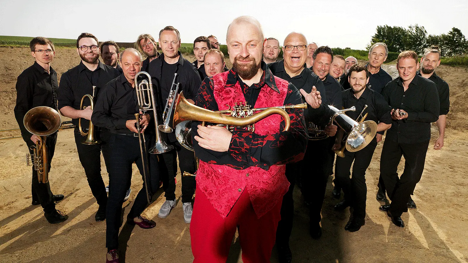 Gruppenfoto der Blasmusik Supergroup im Freien. Im Vordergrund hält eine Person in roter Weste ein Blasinstrument, während zahlreiche Musikerinnen und Musiker in schwarzer Kleidung mit Trompeten, Posaunen, Tuben und weiteren Blechblasinstrumenten dahinter stehen.