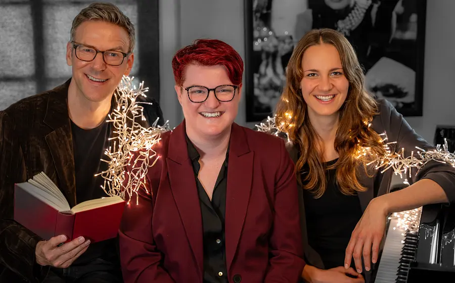 Alexander Netschajew und Duo Key mit drei Personen, Alexander Netschajew hält ein rotes Buch, rechts sitzt eine Pianistin am Klavier. In der Mitte eine Person in dunkelrotem Blazer, eine Lichterkette verläuft durch das Bild, im Hintergrund hängt ein schwarz-weiß Bild
