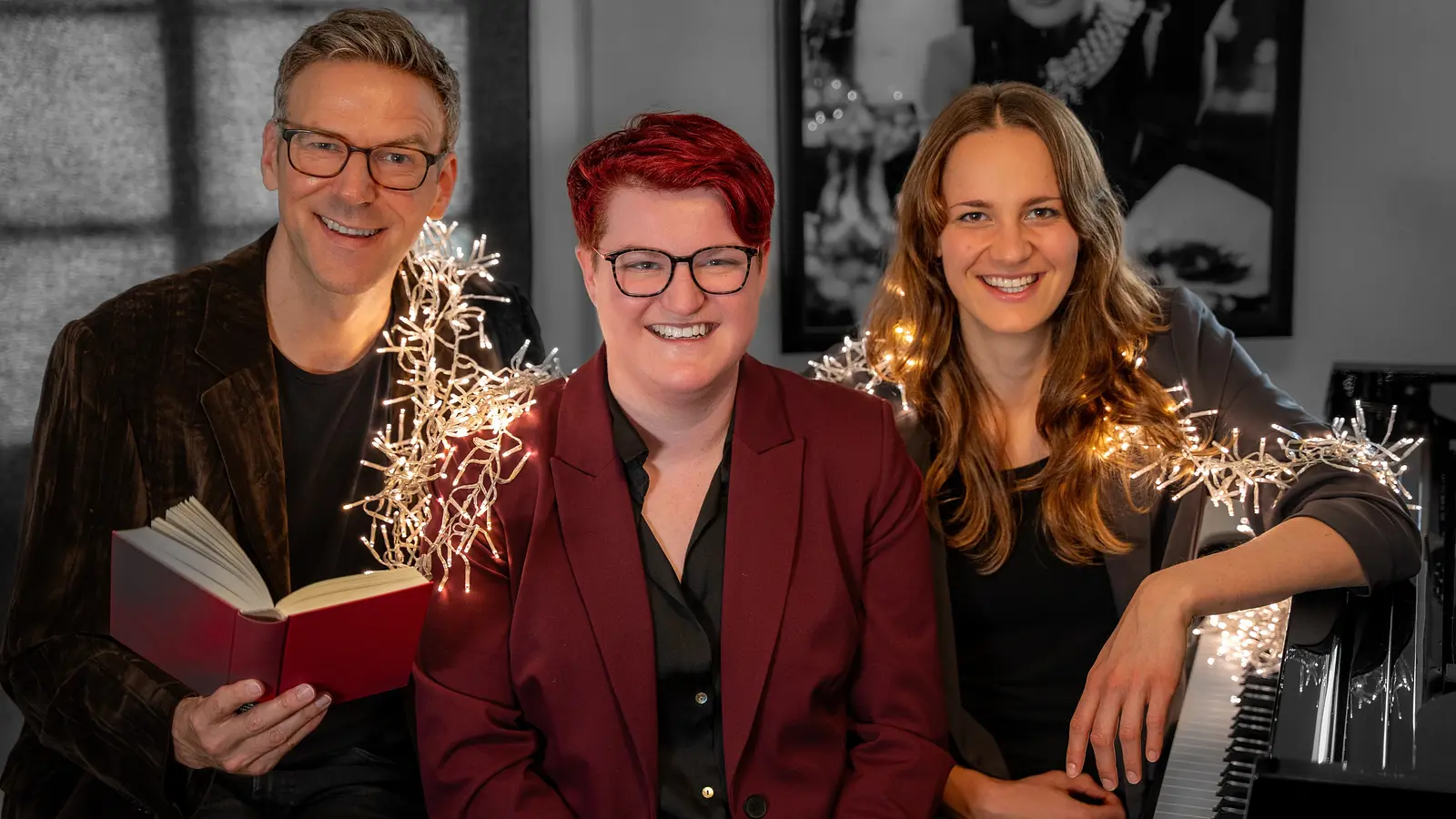Alexander Netschajew und Duo Key mit drei Personen, Alexander Netschajew hält ein rotes Buch, rechts sitzt eine Pianistin am Klavier. In der Mitte eine Person in dunkelrotem Blazer, eine Lichterkette verläuft durch das Bild, im Hintergrund hängt ein schwarz-weiß Bild